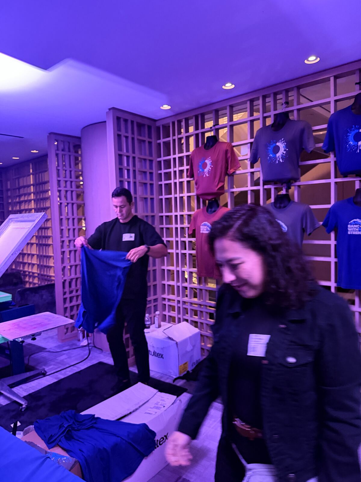 Staff members at an indoor event booth with purple ambient lighting handling blue t-shirts and merchandise